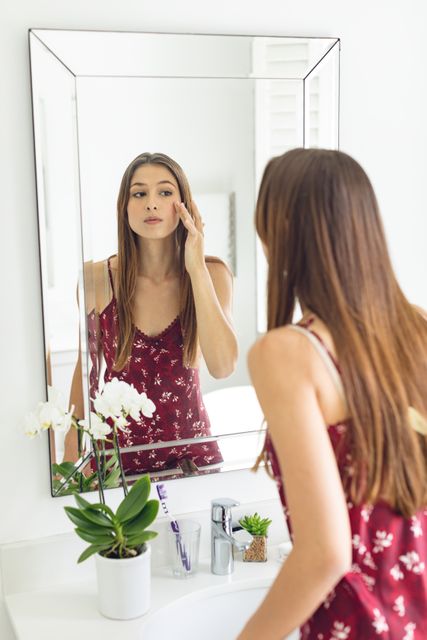 Young woman in casual wear examining her face in bathroom mirror. Perfect for articles or advertisements related to skincare, self-care practices, daily beauty routines, home lifestyle, or personal care products.