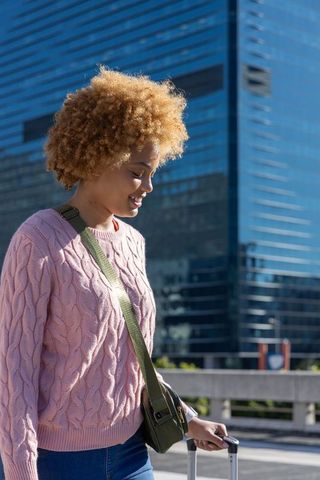 African american woman walking on urban street wearing pink sweater pulling suitcase
