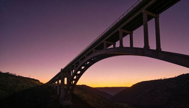 Spanning concrete arch viaduct glowing at twilight over canyon and silhouetted hills