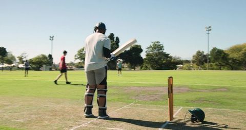 African american cricketer preparing at crease on sunny pitch with team in background