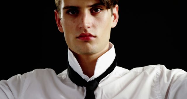 Young man wearing a white dress shirt and a black tie against a black background. He is loosening his tie and looking serious. This image may be used to represent work stress, fashion, formal wear, or professional scenarios in advertisements, websites, or marketing materials.