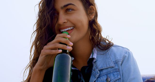 Image of a young woman with brown hair smiling while holding a bottle of beer outdoors. She is wearing a denim jacket and appears to be enjoying a casual and relaxed moment. Useful for articles and advertisements related to youth culture, social gatherings, outdoor leisure, beverages, and lifestyle. Ideal for use in promotional materials and websites targeting a youthful demographic and premises promoting alcohol and social events.