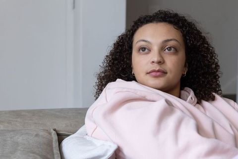 African american woman sitting on beige sofa wearing pink hoodie looking contemplative