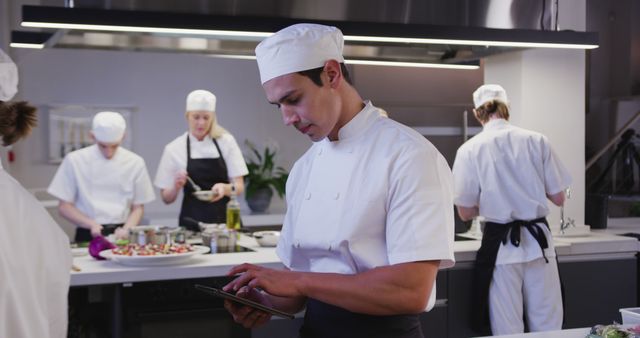 Professional chef using tablet to manage kitchen workflow in a modern restaurant setting. Other kitchen staff in background. Ideal for themes related to culinary arts, digital technology in the kitchen, team collaboration, and modern restaurant operations.