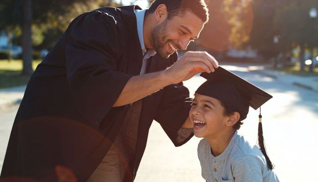 Father in graduation gown placing mortarboard on son's head celebrating proud milestone