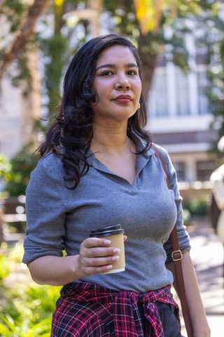 Confident woman standing in sunlit courtyard holding coffee cup and wearing plaid shirt