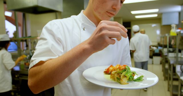 Young chef garnishing a dish with fresh herbs in a busy professional kitchen. Image can be used for promoting culinary schools, restaurant businesses, food blogs, and cooking classes.