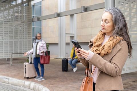 Mature woman checking phone while waiting for rideshare pickup, luggage on curb in city
