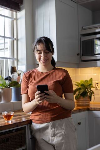 Asian woman smiling and scrolling smartphone while leaning on modern kitchen counter with tea