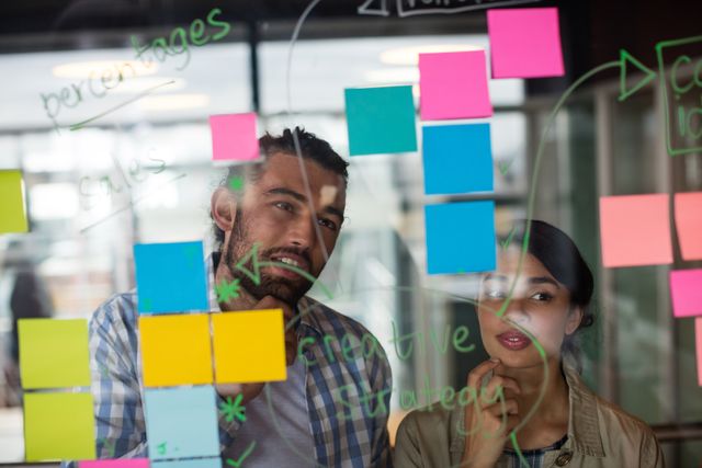 Two executives are brainstorming and discussing ideas using colorful sticky notes on a glass wall in a modern office. This image can be used for business presentations, articles on teamwork and collaboration, creative strategy planning, and project management resources.