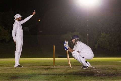 Night cricket scene batsman crouching in crease holding bat while umpire raising finger under floodl