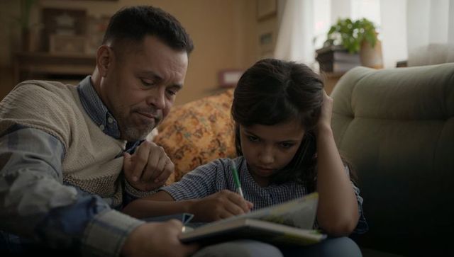 Father guiding daughter writing in notebook on couch sharing focused homework moment