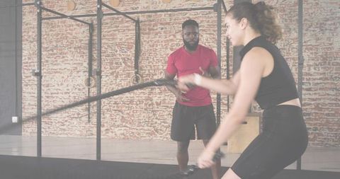 Female athlete using battle ropes while trainer coaching in industrial brick gym