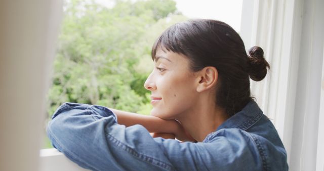 Woman is resting her arms on windowsill, looking outside. Temporary moment conveys serenity and introspection. Suitable for themes of leisure, relaxation, mental health, wellbeing, nature, self-reflection.