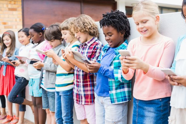 Diverse group of kids standing in a row outdoors, each using mobile phones and tablets. Ideal for illustrating modern education, technology use among children, digital learning, and social interaction in school environments.