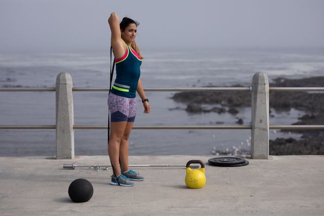 Side view of a strong Caucasian woman with long dark hair wearing sportswear exercising outdoors by the seaside on a sunny day, strength training using black rubber tape, ball and kettlebell next to her.