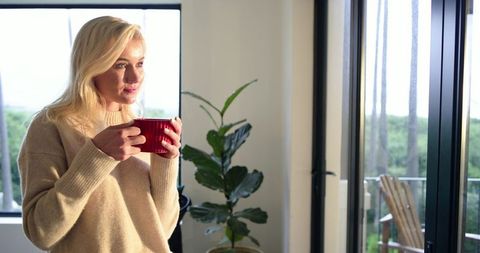 Blonde woman holding red mug, gazing toward balcony in sunlit cozy minimalist living room