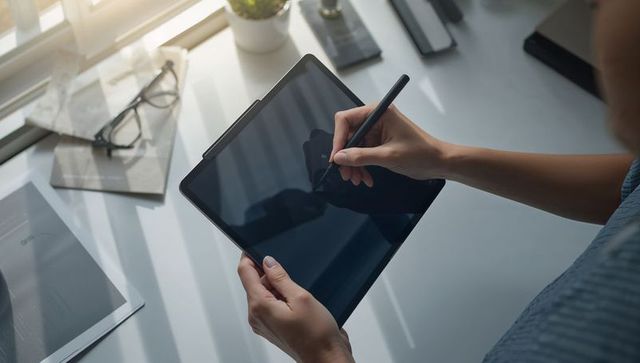 Woman holding tablet sketching with stylus at sunlit desk near window, creative work flow