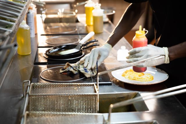 African American male chef cleaning a frying pan in a busy restaurant kitchen. The chef is wearing gloves and working diligently to maintain hygiene. This image is ideal for use in articles or advertisements related to the food industry, restaurant operations, culinary arts, and kitchen hygiene.