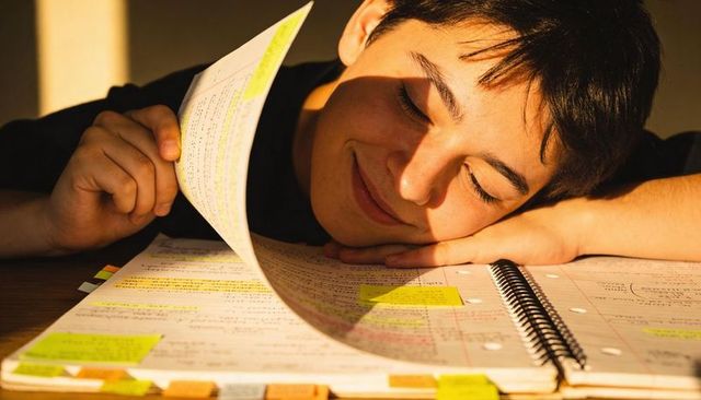 Teen resting and smiling on desk while flipping highlighted notes in warm sunlight