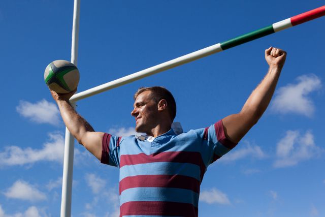 Low angle view of smiling rugby player holding ball with arms raised by goal post against blue sky