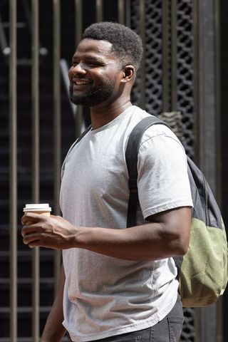 African man smiling and holding coffee cup while carrying backpack at urban gate