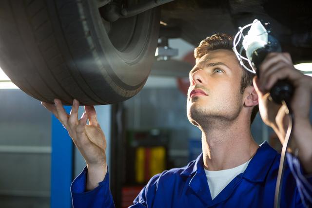 This image shows a mechanic in a blue uniform inspecting a car tire using a flashlight in a repair garage. The scene emphasizes attention to detail and professional service. It can be used for automotive repair advertisements, websites, mechanic training materials, and articles about vehicle maintenance.