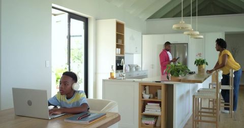African american child studying on laptop while parents chatting in bright modern kitchen