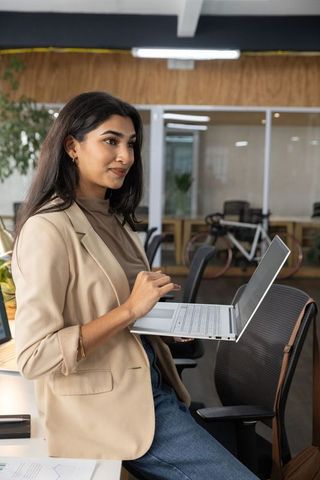 Indian professional leaning on desk using laptop in modern glass-walled coworking office