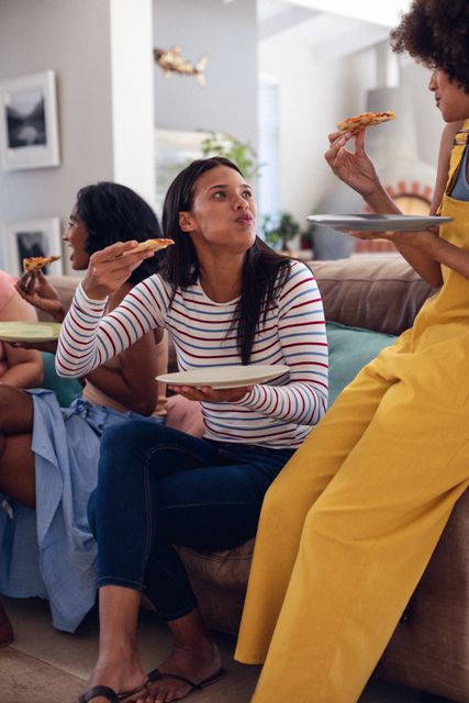 Biracial young female friends enjoying pizza slices while spending leisure time at home, copy space. Food, unaltered, friendship, togetherness, social gathering, enjoyment and weekend activities.