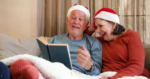 Senior Couple Reading Together on Couch Wearing Santa Hats Celebrating Cozy Holiday