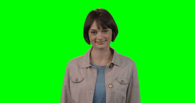Young woman is smiling while posing in a studio against a green screen backdrop. She is wearing casual attire, including a light-colored jacket and a blue shirt. The green screen makes it easy to change the background, making this image highly versatile for use in various projects like advertisements, promotional materials, or creative designs involving added effects.