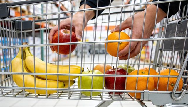 Hands placing apple and orange into shopping cart holding bananas and fresh produce