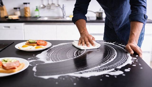 Man wiping soapy black kitchen countertop with white cloth, breakfast plates nearby