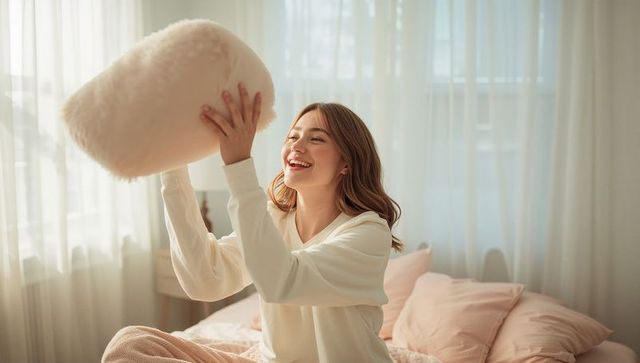 Smiling woman tossing fluffy pillow on cozy pastel bed with sheer curtains and sunlight