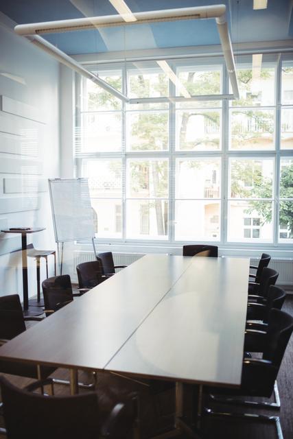 This image depicts an empty modern business meeting room with a large conference table and chairs arranged around it. The room features large windows allowing natural light to flood in, creating a bright and inviting atmosphere. The clean and contemporary design makes it suitable for corporate presentations, team meetings, and professional discussions. Ideal for use in business-related content, office environment promotions, and corporate training materials.