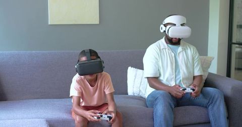 African American Father and Son Wearing VR Headsets Playing Video Games on Home Couch