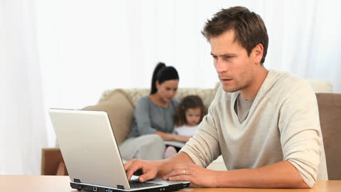 Man Focusing on Laptop While Family Interacts in Living Room