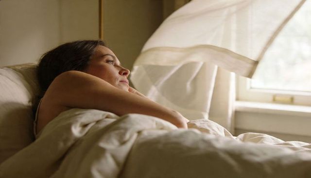 Woman resting in morning light on bed with sheer curtain and neutral bedding