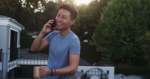 Happy Asian Man Talking on Smartphone while Relaxing on Balcony