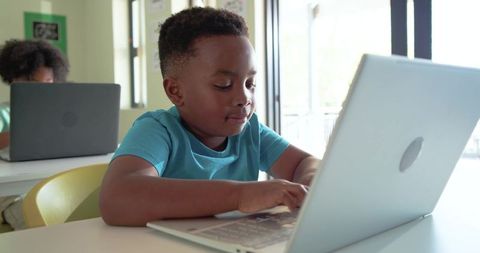 Young African American boy typing on laptop in bright classroom studying technology skills