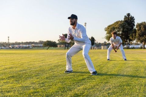 Cricket teammates fielding on sunlit outfield at sunset wearing whites and black cap