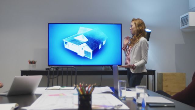 Businesswoman showcasing 3D architectural model on a monitor during a conference in a modern office setting. Ideal for depicting collaborative work environments, innovative technology in business processes, architectural presentations, and professional settings focused on design and innovation.