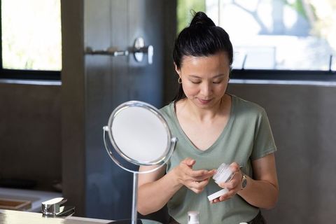 Mature asian woman applying moisturizer at bathroom vanity mirror, daily skincare routine