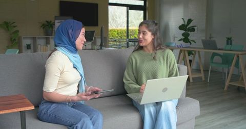 Woman wearing hijab and colleague collaborating on laptop and tablet in modern workspace