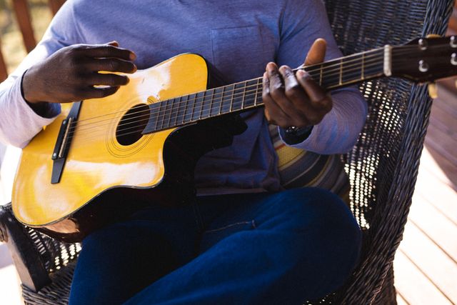 Midsection of african american senior man playing guitar while sitting on chair in log cabin. Unaltered, retirement, vacation, solitude, music, hand, relaxation, hobby and leisure concept.