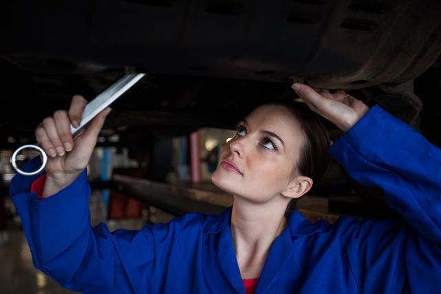 Female mechanic in blue uniform working under a car in a repair garage, using a wrench. Ideal for depicting women in skilled trades, automotive industry, and gender diversity in the workplace. Can be used in articles, advertisements, and educational materials promoting car maintenance and repair services.