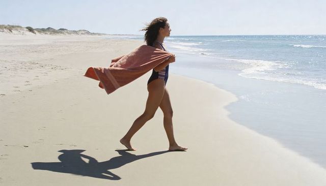 Woman walking along empty sandy beach with towel in one-piece swimsuit by ocean waves