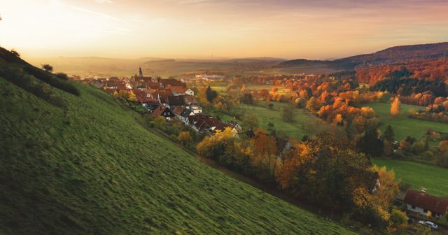 Small village surrounded by picturesque countryside and rolling hills during sunrise in autumn. Can be used for travel, nature, or seasonal content showcasing tranquility and scenic beauty.