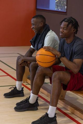 Two african american men sitting on gym bench holding basketball laughing together